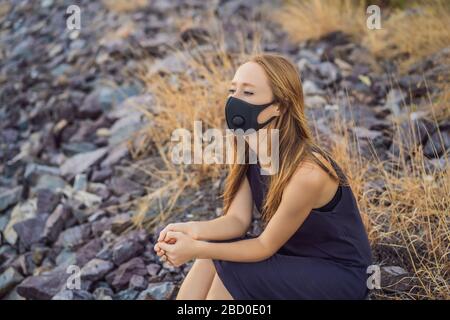 Selbstisolierung in der Natur. Junge Frau in modischer schwarzer medizinischer Maske mit Filter am Fluss sitzend. Coronavirus 2019-ncov Epidemiekonzept Stockfoto