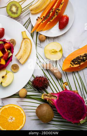 Obere Ansicht der Platten mit geschnittenen Früchten. Auswahl an verschiedenen exotischen Früchten mit tropischen Blättern auf Marmorgrund. Köstlicher Snack, Vitaminheilt Stockfoto