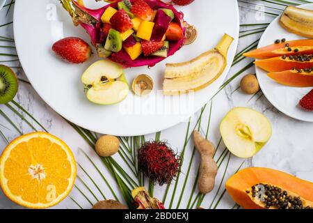 Ein Set exotischer reifer, saftiger, saisonaler tropischer Früchte verteilt sich auf Platte und Tisch. Draufsicht auf geschnittene Früchte und tropische Blätter. Flat Lay Summer com Stockfoto