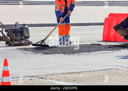 Der Straßenbauer verteilt einen Teil des Asphalts gleichmäßig mit einer Holzebene auf einem Teil der Straße, der mit einem orangefarbenen Kegel und einem roten Zaun eingezäunt ist. Stockfoto