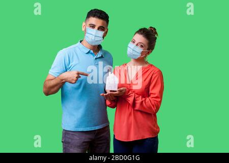 Portrait des glücklichen Paares mit chirurgischer medizinischer Maske, die mit Glück auf die Kamera blickt, Mann, der in den Händen der Frau auf das Haus zeigt. Bleiben Sie zu Hause, bleiben Sie sicher auf qu Stockfoto