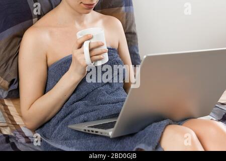 Freiberuflerin arbeitet mit blauer Bettwäsche an ihrem Laptop mit einer Tasse Tee im Bett. Konzept des Heimbüros. Frau entspannt sich nach dem Bad, in das sie eingedeckt ist Stockfoto