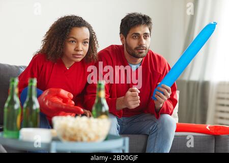 Portrait eines modernen Mixed-Race-Paares, das Sport zu Hause im Fernsehen verfolgt und sich für intensives Match jubelt, während es rote Teamuniformen trägt, kopiere Platz Stockfoto