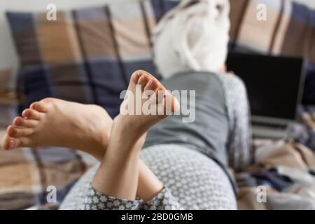 Nahaufnahme von Womanen gekreuzte Beine. Rückansicht auf eine liegende junge Frau im Bett und mit einem Laptop. Konzept Freizeit und Wochenende. Stockfoto