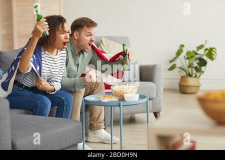 Seitenansicht des jungen Paares, das Sportmatches zu Hause im Fernsehen beobachtet und emotional jubelt, während man amerikanische Flagge trägt, kopiere Raum Stockfoto
