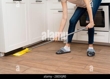 Eine junge Frau wäscht Holzböden aus einem Laminat in einer hellen Küche. Das Mädchen wäscht Staub und Schmutz unter dem Küchenschrank. Stockfoto