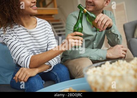 Zugeschnittenes Porträt eines modernen Mixed-Race-Paares, das zu Hause Fernsehen und Bierflaschen klinken, während es auf dem Sofa in einer gemütlichen Wohnung sitzt Stockfoto