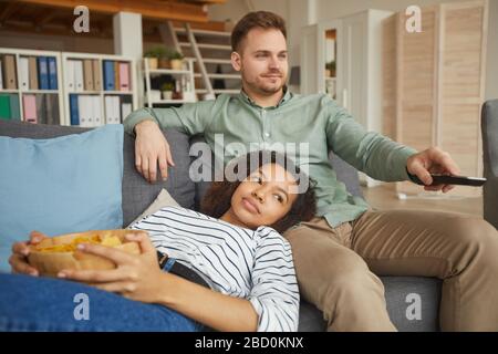 Portrait eines jungen Mixed-Race-Paares, das zu Hause Fernsehen und Snacks isst, während es auf einem bequemen Sofa und einem Kopierraum entspannen kann Stockfoto