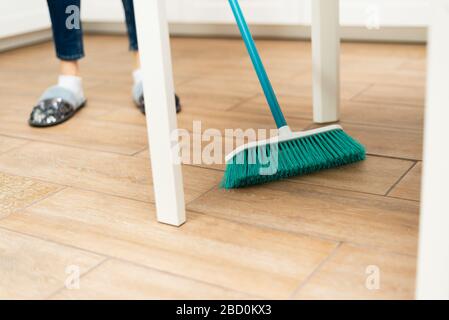 Junge Frau fegt Laminatboden in einer hellen Küche. Das Mädchen fegt Staub und Schmutz unter dem weißen Küchentisch. Stockfoto