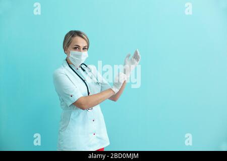 Frau Doktor oder Krankenschwester, die eine Maske, einen Gown und ein Stethoskop tragen und auf blauem Hintergrund Operationshandschuhe aufsetzen. Stockfoto