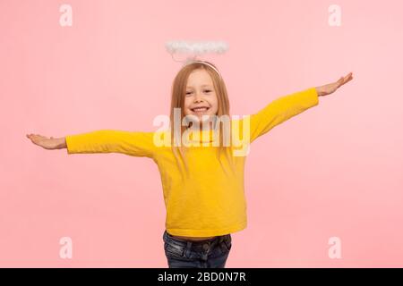 Schönes Engelskleines Mädchen mit Halo über dem Kopf, das die Hände hebt, als ob es fliegt und mit tollem Lächeln auf die Kamera blickt, Konzept eines freundlichen, gehorsamen Kindes. Stockfoto