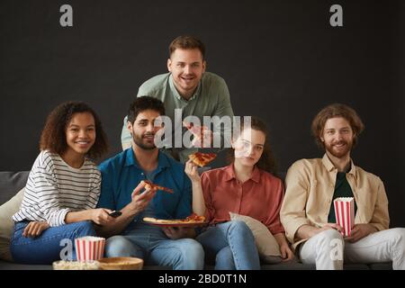 Große Gruppe von Freunden, die Pizza und Snacks essen, während sie die Party zu Hause auf einem großen Sofa vor schwarzem Hintergrund genießen und alle auf die Kamera schauen, Co Stockfoto
