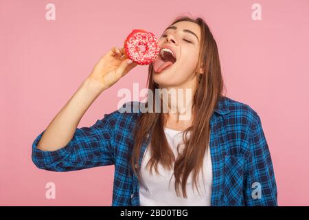 Hübsches Mädchen im karierten Hemd, das mit geschlossenen Augen steht und süße Donuts leckt, träumt davon, leckeren Donut zu beißen, und kämpft mit der Versuchung, suga zu essen Stockfoto