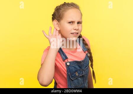 Was? Ich kann es nicht hören! Porträt eines neugierigen, aufmerksamen kleinen Mädchens in Denim-Overalls, das die Hand in der Nähe des Ohrs hält und aufmerksam auf das hört, was Sie sagen Stockfoto