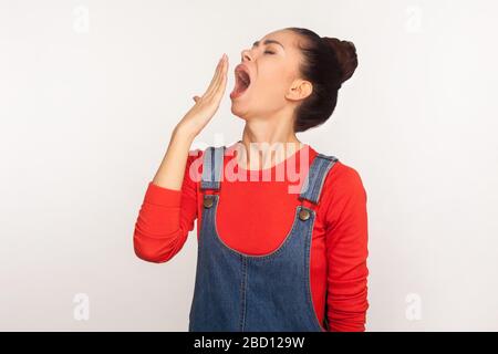 Schlaf erforderlich. Portrait eines erschöpften Ermüdungsmädchens mit Haarbun in Denim-Overalls gähnend und den Mund mit der Hand bedeckend, faul, leidenswert insomni Stockfoto