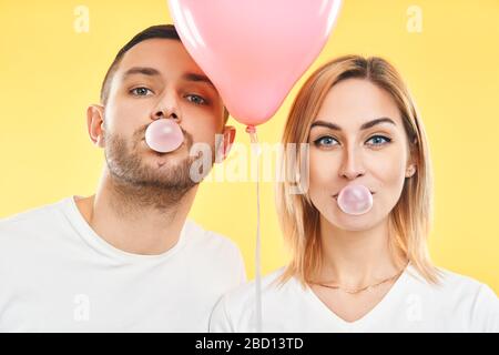 Junges Paar bläst Blase mit Kaugummi und hält rosa Luftballon auf gelbem Hintergrund Stockfoto