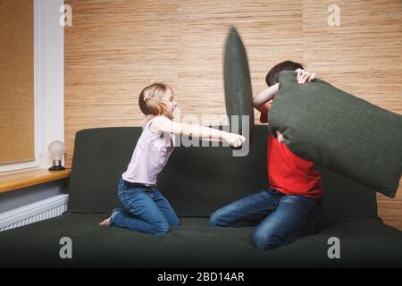 Geschwister, die verrückt werden, steckten zu Hause in Selbstisolation fest. Kinder kämpfen mit Kopfkissen auf einer Couch. Quarantäne und Sperrung der Schutzmaßnahmen erneut Stockfoto