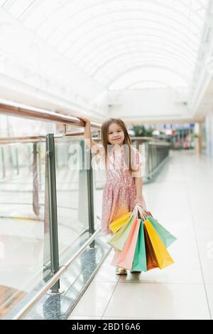 Kinder, die Einkaufstaschen halten. Kleines Mädchen in rosafarbenem Kleid, das durch das Schaufenster blickt. Ein lächelndes lachendes Mädchen in einem rosafarbenen Kleid mit mehrfarbigen Taschen in den Händen läuft um das Einkaufszentrum und schaut auf die Schaufenster. Stockfoto