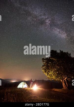 Weibliche Wanderer, die sich in der Sommernacht auf dem Campen in den Bergen neben dem Lagerfeuer, dem leuchtenden Touristenzelt und dem großen Baum entspannen. Junge Frau, die einen Blick auf den Nachthimmel voller Sterne und Milchstraße genießt. Stockfoto