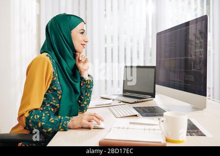 Lächelnde junge muslimische Frau in traditionellem Kleid genießt es, an einem neuen Projekt für IT-Unternehmen zu arbeiten Stockfoto