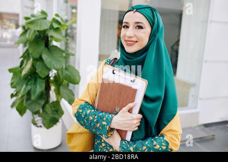 Porträt einer lächelnden muslimischen jungen Geschäftsfrau mit Aktenordner auf der Straße und Blick auf die Kamera Stockfoto