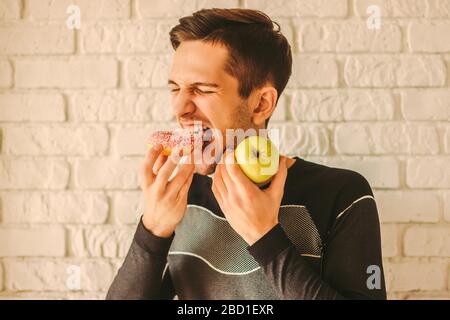 Junge attraktive Sportmann essen Donut Kuchen und halten Apfel in der Hand. Gieriger Mann Fitness-Trainer Wahl zwischen süßen Snack oder gesunde Früchte. Fitne Stockfoto