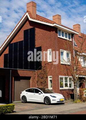 Tesla Model 3 Electric EV Car Charging zu Hause vor dem modernen Low Energy House mit Solarmodulen. Umweltkonzept Green Energy. Stockfoto