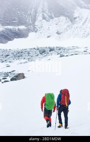 Rückansicht der männlichen Bergsteiger mit Rucksack, der durch den Schnee geht und zum Gipfel führt. Reisende, die in verschneiten Bergen wandern. Reisekonzept, Alpinismus und aktive Freizeit. Stockfoto