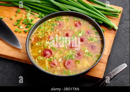 Trockene Erbsensuppe mit Fleischwürsten, Kartoffel, grünen Zwiebeln und Sesam. Gelbe Pea-Suppe in schwarzer Schüssel mit dunklem Schieferhintergrund. Stockfoto