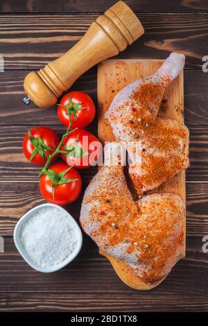 Rohe Hähnchenbeine auf einem Schneidebrett auf einem Holztisch. Mariniertes Hähnchenbein mit Gewürzen und Tomaten. Vertikales Foto Stockfoto