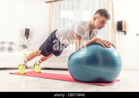 Arbeiten Sie zu Hause von einem jungen Mann, der eine Plankposition mit einem blauen Fit Ball und einer roten Matte in einem Wohnzimmer hält. Stockfoto