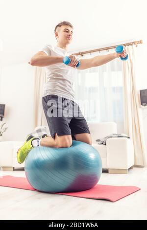 Sportlicher kaukasischer junger Mann, der auf einem Fitnessball kniet, der aufrecht steht, mit einem Arm, der mit Hanteln in den Händen ausgestreckt ist. Stockfoto