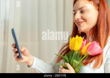 Wunderschönes glückliches Mädchen lächelt, hält mit Frühlings-Tulpen-Blumenstrauß und nimmt selfie-foto im Zimmer auf. Frauentag 8. März Stockfoto
