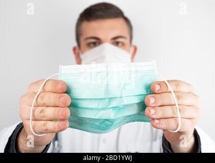 Kaukasischer Mann posiert als Arzt in weißer Gesamt- oder Gestrüpp, der eine schützende Gesichtsmaske hält. Stockfoto