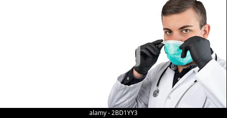 Kaukasischer Mann posiert als Arzt in Weiß insgesamt oder schrubbt mit Stethoskop um einen Hals und schwarze Handschuhe. Aufsetzen einer Gesichtsschutzmaske. Stockfoto