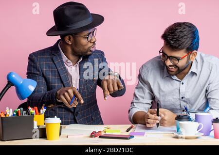 Zwei selbstbewusste Geschäftsleute, die Anzüge tragen, sitzen am Schreibtisch im Büro. Dunkelhäutiger Manager, der Smartphone hält und auf den Bildschirm des Gadgets schaut Stockfoto