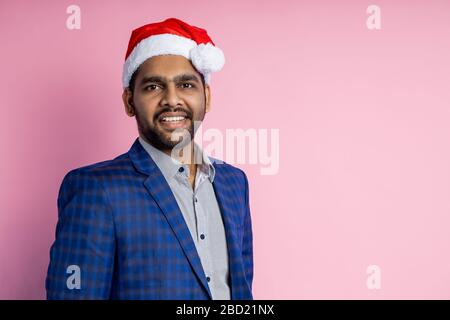 Horizontales Studio-Porträt des jungen, freundlichen indianischen Unshaven, Mitarbeiter, Arbeiter, der Anzug trägt, Santa Hut, Blick auf die Kamera mit ruhigem Lächeln, Isolieren Stockfoto