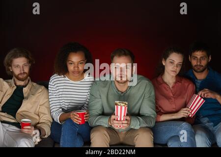 Eine Gruppe von Freunden, die zu Hause Filme ansehen, während sie Snacks und Popcorn essen, die auf einem großen Sofa in einem dunklen Zimmer sitzen Stockfoto