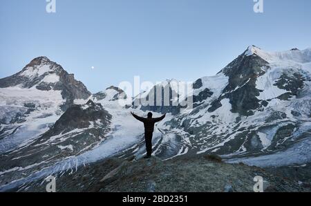 Fröhlicher Reisender, der die Nacht in den Bergen begrüßt und mit dem Rücken zur Kamera steht, mit offenen Händen auf den Gletscher und die hohen schneebedeckten Gipfel der Pennine Alpen blickt Stockfoto