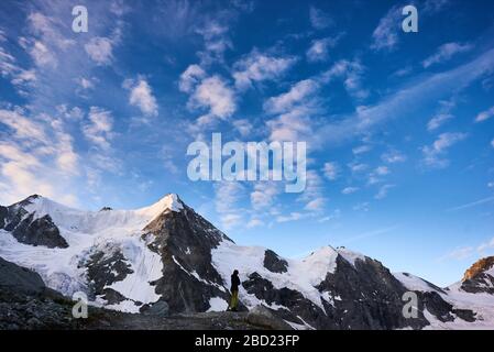 Zinal, Schweiz - 19. Juli 2019: Voller Wanderer, der die fantastische Aussicht auf schneebedeckte Berge unter blauem bewölktem Morgenhimmel bewundert. Konzept des Reisens, Wanderns und Bergsteigens. Stockfoto