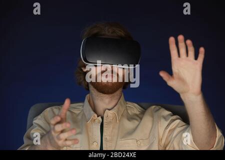 Kopf- und Schulterporträt des modernen bärtigen Mannes, der virtuelle Reality-Ausrüstung trägt, während er im Dunkeln immersives Videospiel oder Film genießt Stockfoto