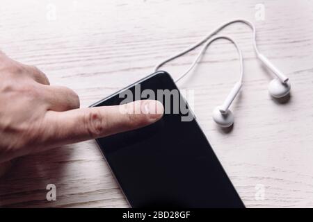 Draufsicht eines Fingers, der ein Smartphone mit Ohrhörern auf weißer Holzoberfläche, Musik- und Podcast-Konzept berührt, Platz für Text kopieren Stockfoto