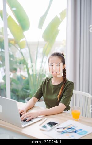 Frau, die zu Hause am Tisch sitzt und mit einem Computerlaptop arbeitet, mit einem fröhlichen Gesicht steht und lächelt mit einem selbstbewussten Lächeln, das Zähne zeigt Stockfoto