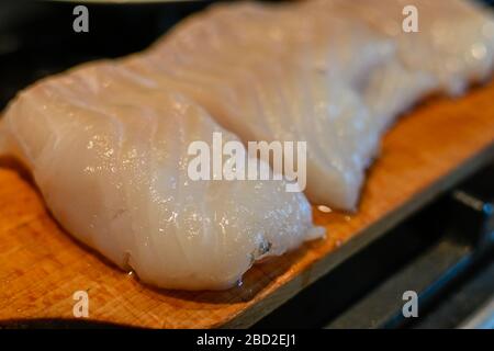 Nahaufnahme eines Kabeljaufilets, das auf eine Holzschneideplatte gelegt und in Portionen geschnitten wurde. Stockfoto