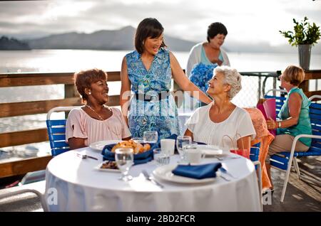 Zwei reife weibliche Freunde, die zum Mittagessen in einem Restaurant am Meer unterwegs sind, lachen mit einem bekannten, der Hallo gesagt hat. Stockfoto