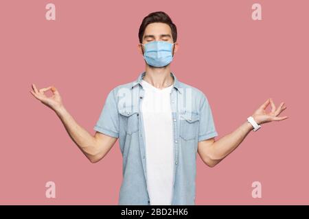 Portrait eines ruhigen, entspannten jungen Mannes mit chirurgischer medizinischer Maske in blauem Hemd, das mit erhobenen Armen steht und Yoga meditiert. Indoor-studi Stockfoto