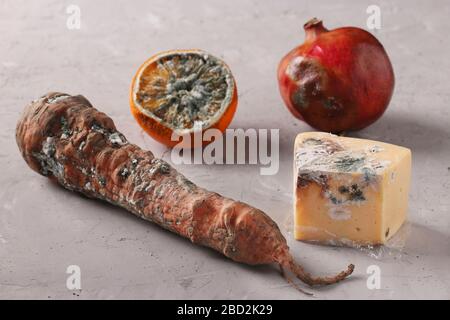 Verdorbene faule Speisen mit Schimmel: Halbe Orange, Granatapfel, Karotten und Hartkäse auf grauem Grund Stockfoto