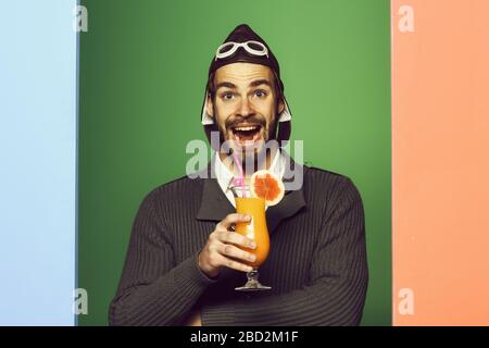 Hübsche bärtige Flieger mit langem Bart auf lächelndem Gesicht, Glas, alkoholischer Cocktail in grauem Strickpullover mit Hut und Brille auf farbenfrohem Studiohintergrund Stockfoto