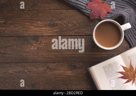 Flatlay-Komposition mit Strickschal, Tasse Kaffee, Buch und gelben Blättern auf Holztisch. Hygge-Stil, gemütliches Herbst- oder Winterferienkonzept Stockfoto