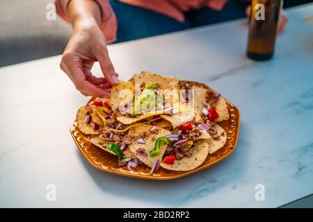 Die Menschen greifen gelbe Mais-Nacho-Chips mit gemahlenem Rindfleisch, Guacamol, geschmolzenem Käse, Paprika und Cilantro-Blättern in Platte auf weißem Stein Stockfoto